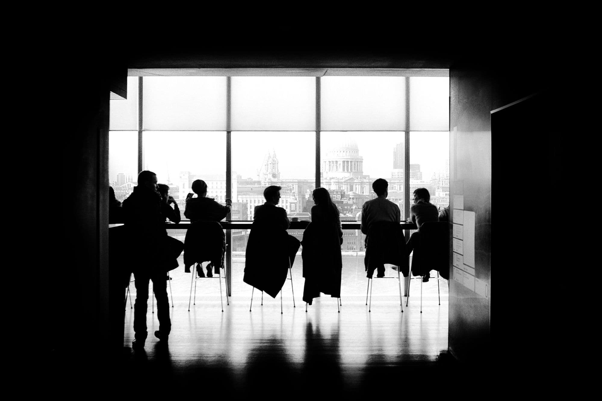 Silhouettes of people in discussion around a long table in front of tall windows overlooking a city