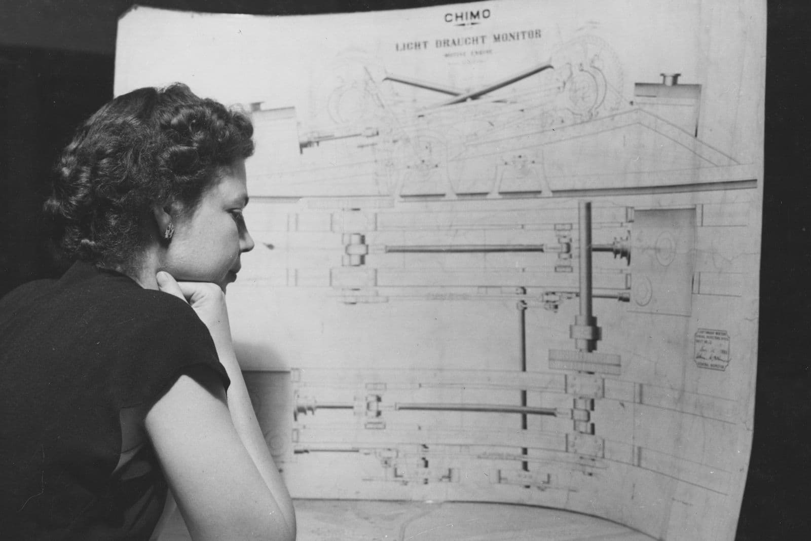 Black and white archival photo of a woman studying a large engineering drawing of a ship
