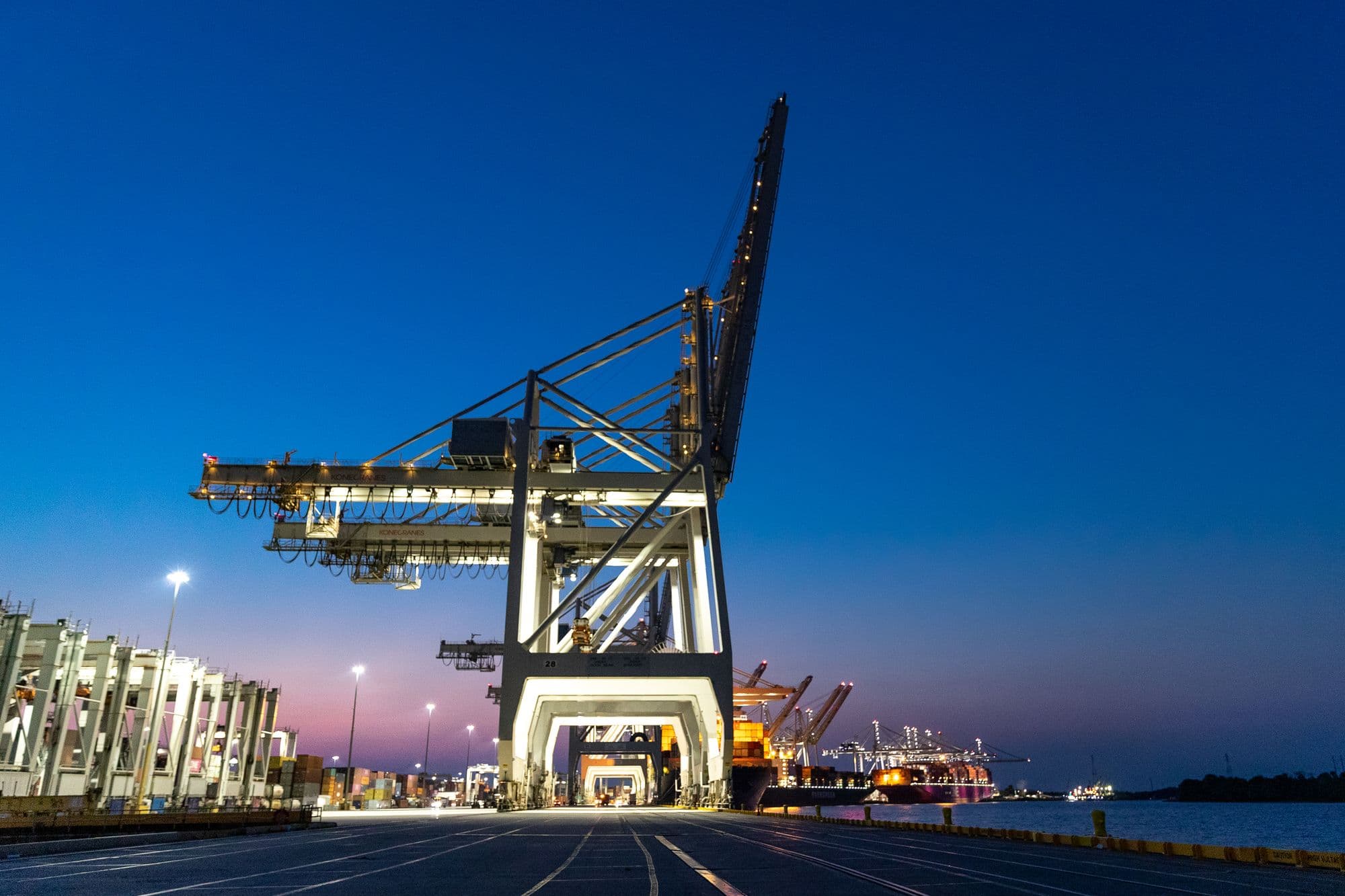 Illuminated quay cranes lining a container terminal at dusk under a deep blue sky