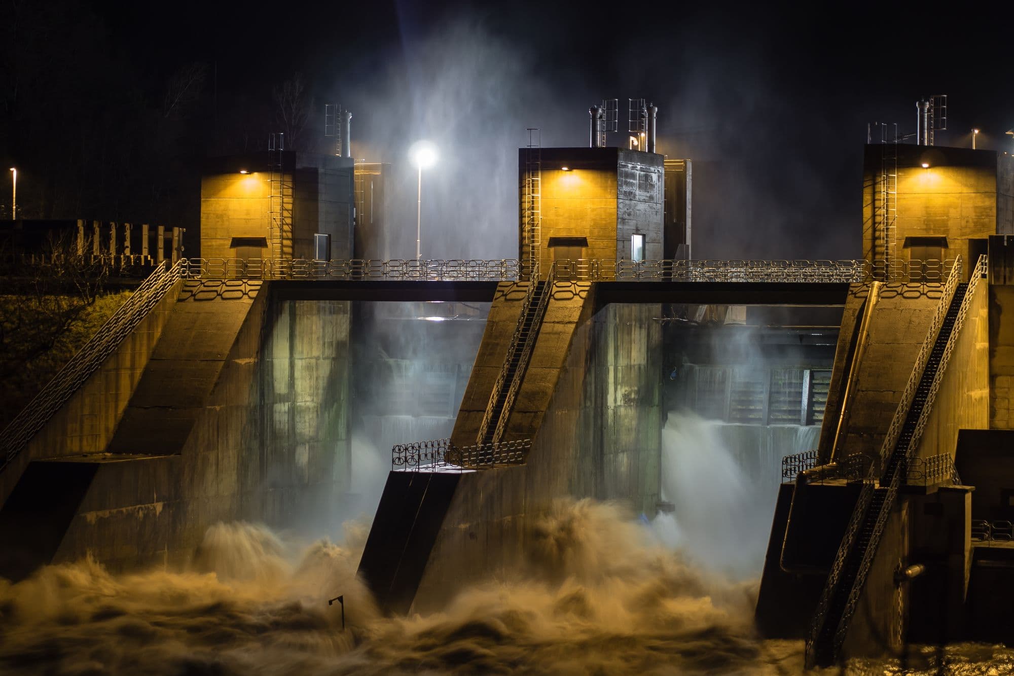 Floodlit dam spillway gates releasing churning water at night