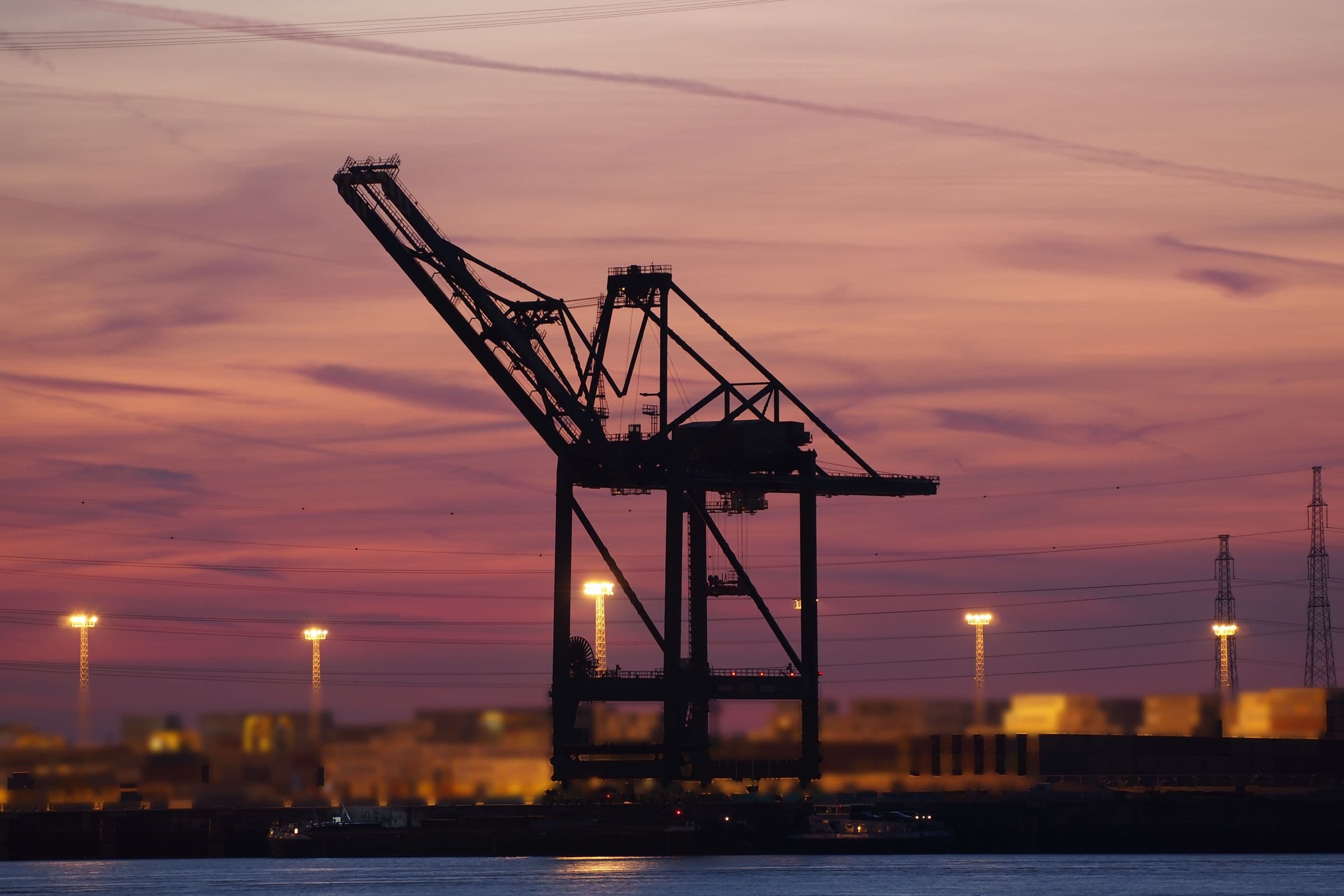Silhouette of a ship-to-shore container crane against a pink and violet sunset sky, with floodlit container stacks along the quay