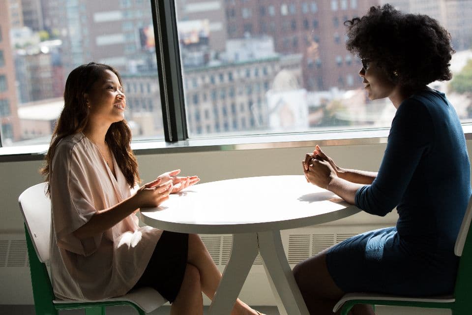 Two women in conversation at a round table beside a large window overlooking city buildings