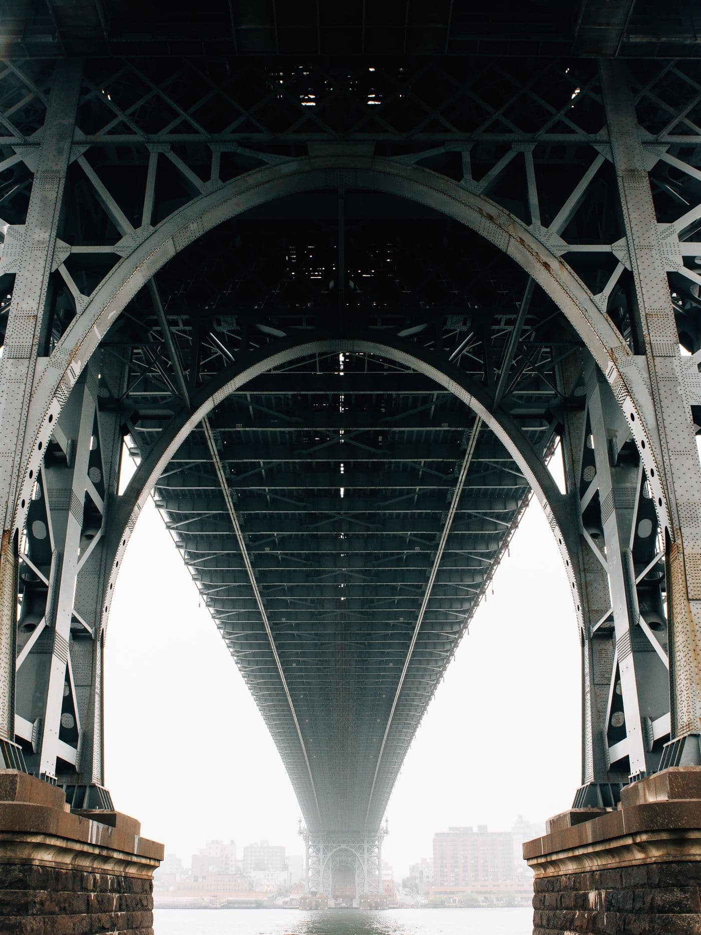 Looking up at the underside of a steel suspension bridge, its deck receding into fog between two towers