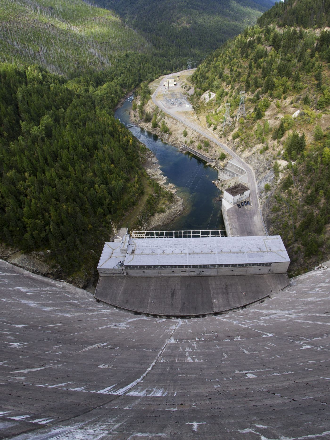 View down the curved face of a concrete dam to the powerhouse and river in a forested valley