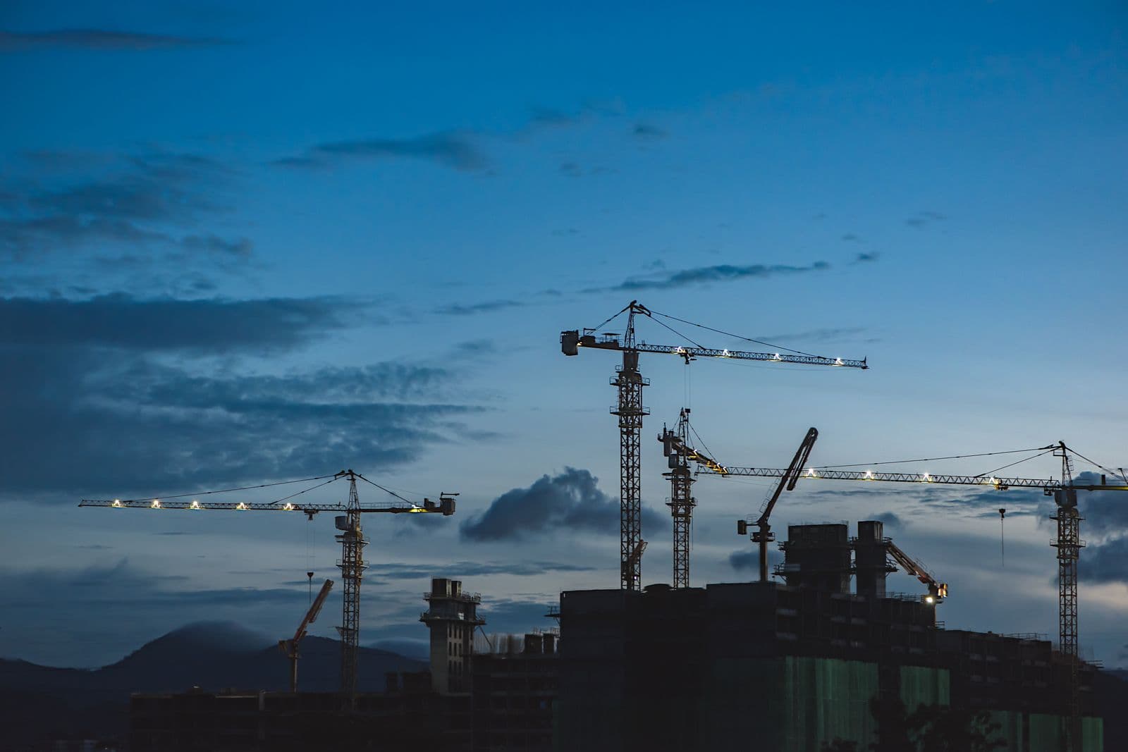 Tower cranes over a building under construction, silhouetted against a blue evening sky